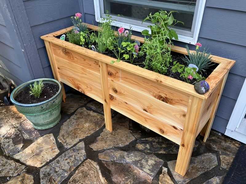 A tall custom cedar planter box on legs, planted with herbs and flowers, on a stone patio